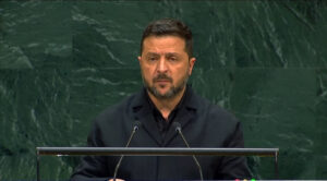 man in a black jacket delivers a speech at a podium with two microphones against a green marble wall